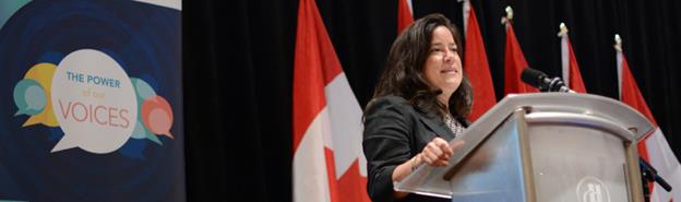 To the left of the photo is a banner featuring multiple layered dialogue bubbles with the middle one stating "the Power of our voices". To the right, Minister Jody Wilson-Raybould speaking at a podium.